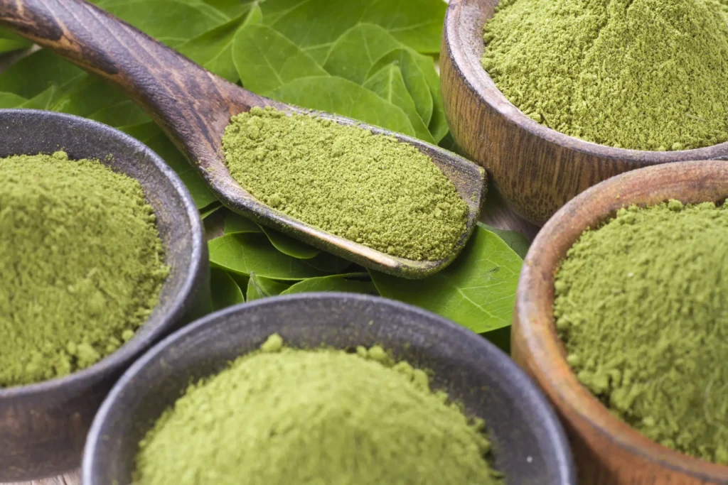 Several bowls of green kratom powder with a wooden spoon and fresh green leaves