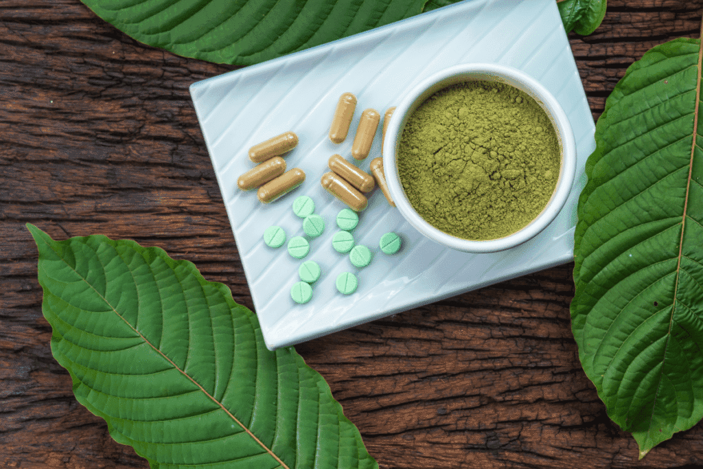 Kratom capsules and green tablets on a white plate beside a bowl of green kratom powder, with kratom leaves on dark wood