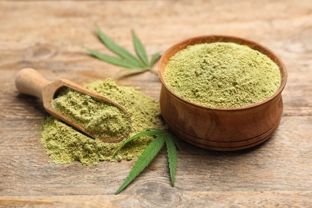 A wooden bowl of green kratom powder with a wooden scoop and fresh leaves on a rustic wooden table
