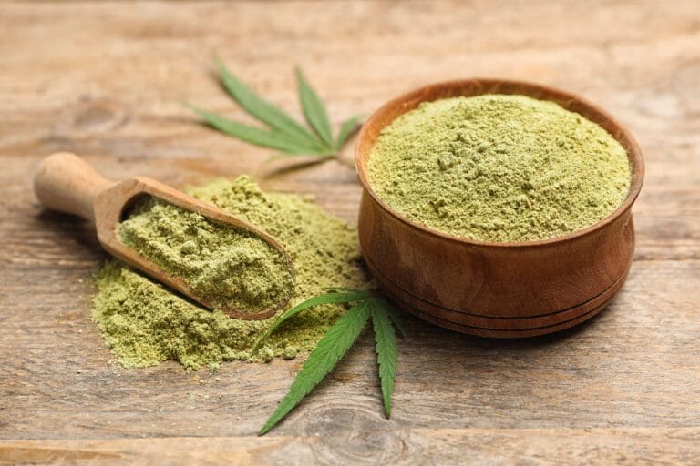 A wooden bowl of green kratom powder with a wooden scoop and fresh leaves on a rustic wooden table