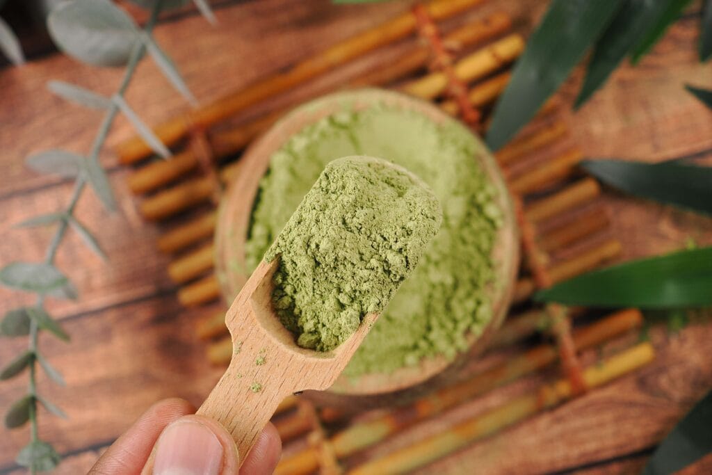 A hand holding a wooden spoon heaped with green kratom powder above a bamboo mat