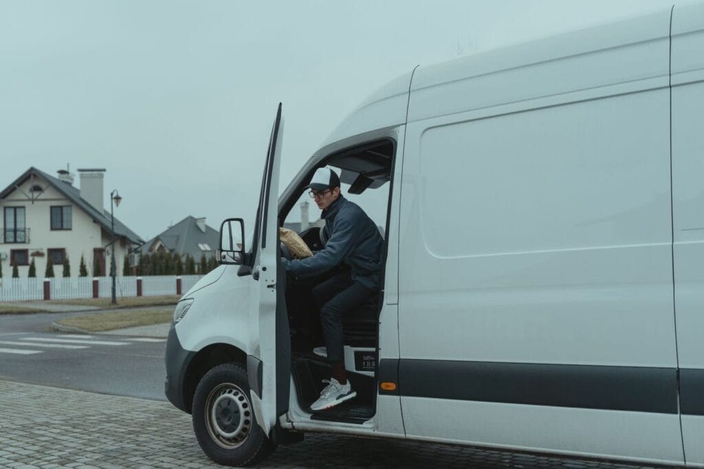 A driver seated in the open door of a white delivery van parked on a quiet residential street