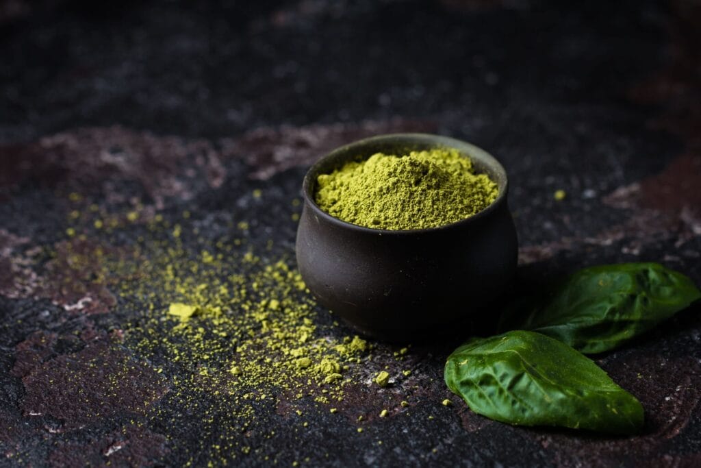 Green kratom powder in a small black bowl with fresh leaves on a dark surface