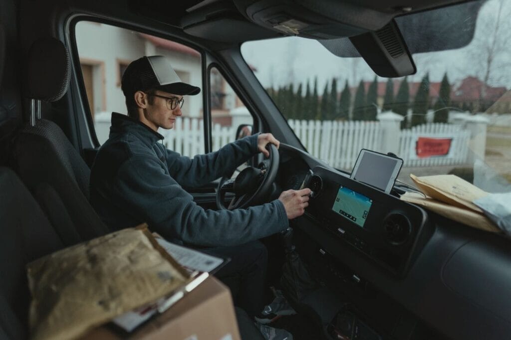 A courier at the wheel of a delivery van with parcels beside them and a route showing on the dashboard screen